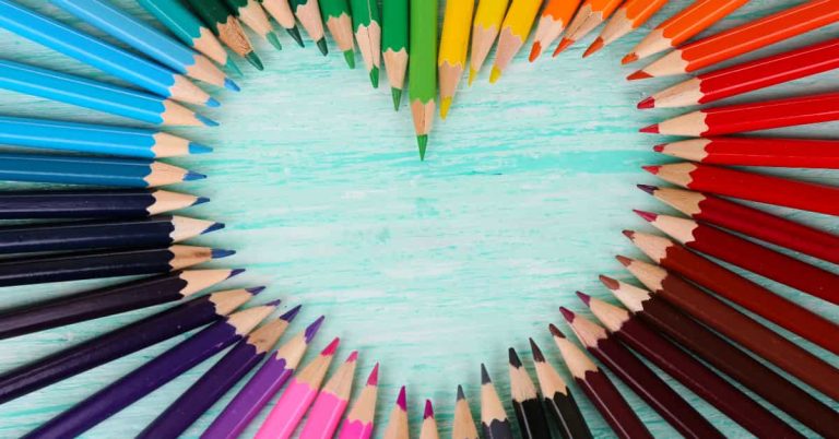 A rainbow selection of coloured pencils, arranged with their points forming a heart shape on a blue wood background